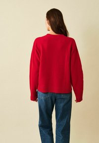 Kvinna med långt brunt hår, klädd i en röd tröja och blå jeans, vänd bort från kameran mot en enfärgad beige bakgrund.