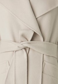 Cappotto avvolgente beige realizzato in morbido pile, con vita cintrata, dotato di un ampio colletto e due tasche frontali. Texture liscia con dettagli minimi.