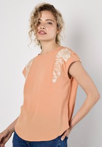 Pfirsichfarbene Bluse mit kurzen Ärmeln und floralem Stickerei an den Schultern. Leichtes Material, abgerundeter Saum und lockerer Schnitt.
