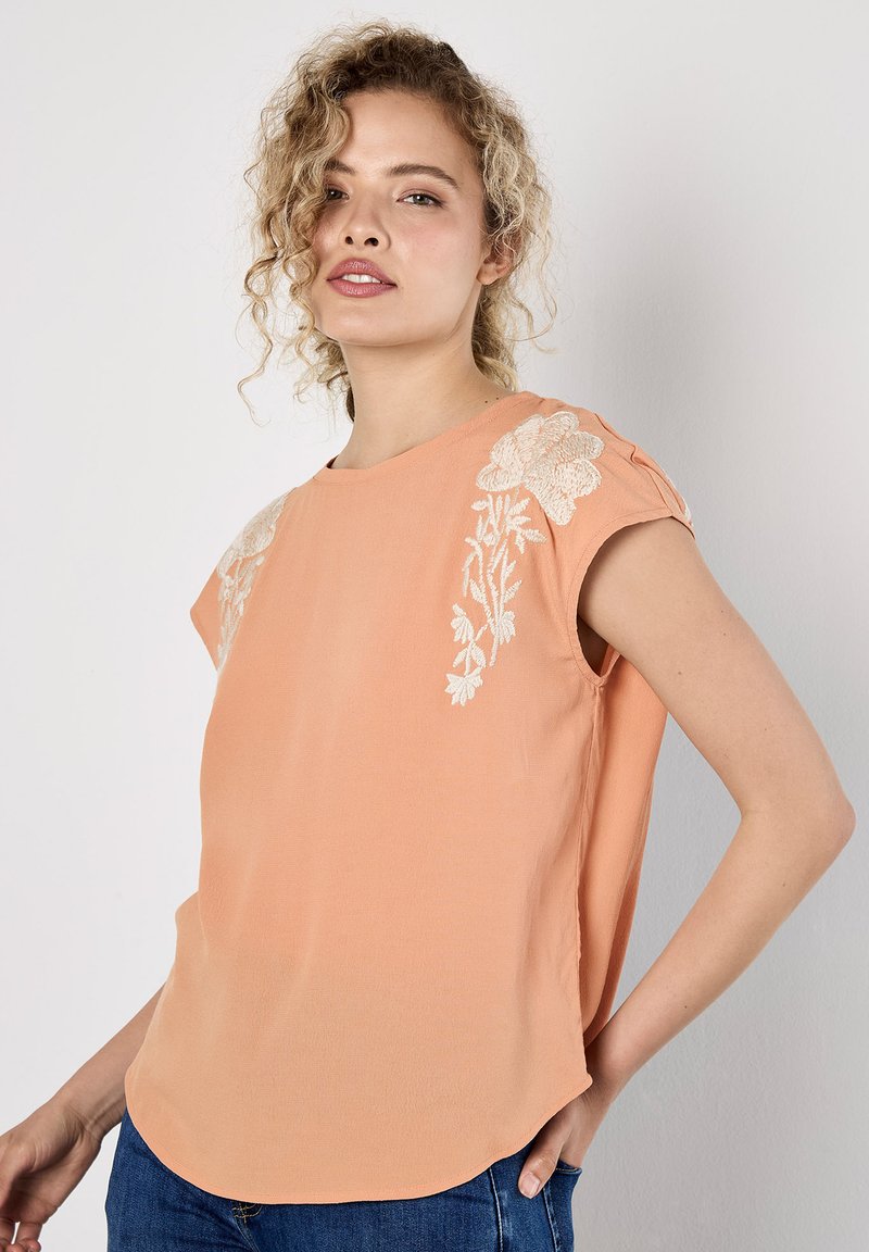 Pfirsichfarbene Bluse mit kurzen Ärmeln und floralem Stickerei an den Schultern. Leichtes Material, abgerundeter Saum und lockerer Schnitt.