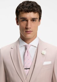 Jeune homme portant un costume rose clair, une chemise blanche, une cravate rose à motif avec pince à cravate, et une épingle à revers en forme de fleur blanche, face à lui.