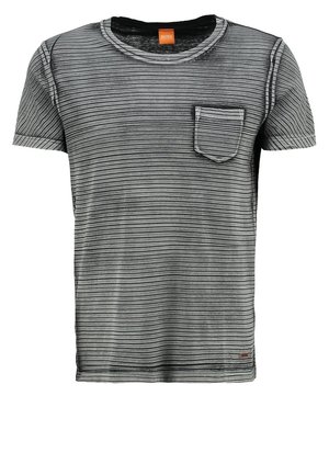 Mężczyzna w pasiastym szarym T-shircie, niebieskich dżinsach i czarnych trampkach z różowym kwiatowym wzorem, trzymający czarne okulary w prawej ręce.