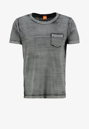 Man die een gestreept grijs T-shirt, blauwe spijkerbroek en zwarte sneakers met een roze bloemmotief draagt, en een zwarte bril in zijn rechterhand vasthoudt.