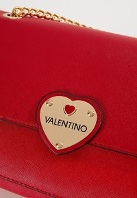 Borsa a tracolla in pelle rossa con una placca dorata a forma di cuore con la parola "VALENTINO" e un piccolo cuore rosso. Include una tracolla a catena. Finitura testurizzata.