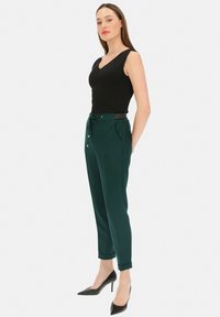 Czarny bezrękawnik noszony z ciemnozielonymi spodniami na sznurek w pasie, z bocznymi kieszeniami i podwiniętymi mankietami. Modelka ma na sobie czarne szpilki.