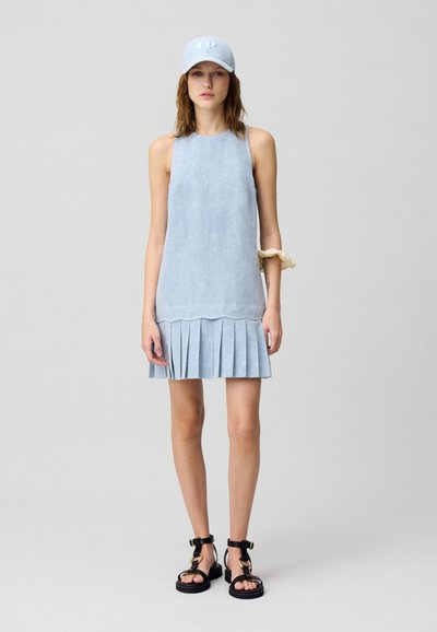 Robe sans manches bleu clair avec un ourlet plissé, confectionnée dans un tissu texturé. Associée à une casquette assortie et des sandales noires avec des accents dorés.
