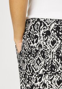 Ręka spoczywa w kieszeni spodni w czarno-białe wzory, na sobie biała bluzka, zbliżenie na bok i teksturę materiału.