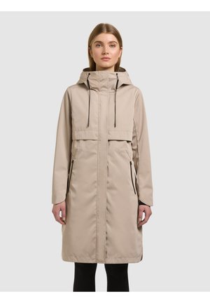 Kobieta w beżowym, długim płaszczu z kapturem, czarnymi ściągaczami i zapinanymi na zamek bocznymi kieszeniami, stojąca na tle jednolitego jasnoszarego tła.