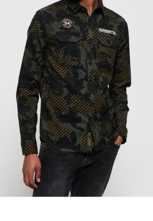 Mann trägt ein grün-schwarzes, camouflagegemustertes Hemd mit Knöpfen, Brusttaschen und dekorativen Abzeichen, kombiniert mit dunklen Jeans.