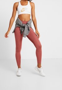Soutien-gorge de sport Nike blanc avec logo, sweat-shirt gris noué autour de la taille, et leggings rouges ajustés, complétés par des chaussures de sport blanches.