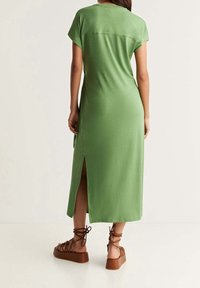 Femme portant une robe midi verte avec une fente sur le côté et des sandales à plateforme marron à lacets, se tenant contre un fond uni.