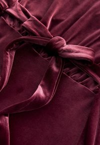 Burgunderfarbenes Samtgewebe mit strukturiertem Finish, ausgestattet mit einem geknoteten Gürtel und gefalteten Details. Durchgehend glatte und weiche Textur.