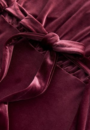 Burgundy sametti, jossa on tekstuuripinta, sisällytään solmittu vyö ja laskostetut yksityiskohdat. Pintakäsittely on sileä ja pehmeä koko matkalla.