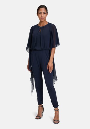 Navy jumpsuit with sheer cape sleeves, keyhole neckline, elastic waistband, and tapered legs. Features strappy high-heeled sandals.