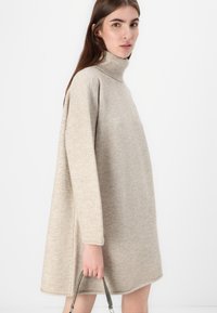 Beige oversized turtleneck kjole i myk, teksturert strikk. Inkluderer lange ermer, senkede skuldre og en rett hem. Håndholdt stropp er synlig.