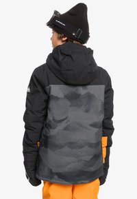 Veste isolée noire et grise avec capuche, présentant un motif texturé au dos, associée à un pantalon orange et un bonnet noir.