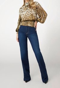 Blouse imprimée à manches bouffantes, associée à un jean évasé bleu foncé avec poches latérales et texture lisse. Fente au niveau de l'ourlet du jean.