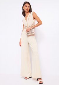 Tuta beige con scollatura a V profonda, dettagli arricciati in vita, design a gamba larga, abbinata a una piccola borsa a tracolla decorata.