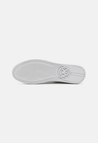 Semelle de chaussure blanche avec motif adhérent texturé et un logo circulaire près du talon sur un fond clair uni.