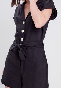 Romper en lin noir avec des manches courtes, fermeture à boutons devant et deux poches latérales. Doté d'une ceinture à nouer à la taille pour un ajustement réglable.