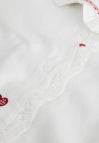 Fehér szövet fodros szegéllyel és kis kerek fehér gombokkal, a gallér szélénél piros hímzett szívekkel díszítve.