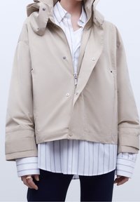Giacca cappuccio beige con zip argentata e bottoni a pressione, indossata sopra una camicia bianca a maniche lunghe con sottili righe verticali, abbinata a pantaloni scuri.