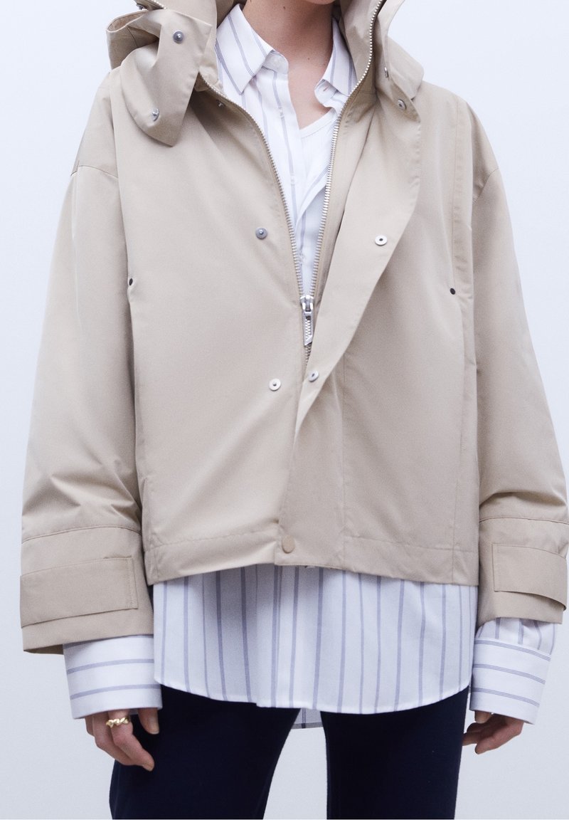 Giacca cappuccio beige con zip argentata e bottoni a pressione, indossata sopra una camicia bianca a maniche lunghe con sottili righe verticali, abbinata a pantaloni scuri.