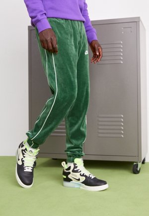 Person mit grüner Velour-Nike-Jogginghose, schwarzen und weißen Nike High-Top-Turnschuhen mit neon-grünen Schnürsenkeln, steht in der Nähe eines grauen Spinds.