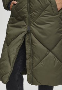 Polstret olivenfarget frakk med kviltet mønster, med en frontglidelås og sidelommer. Stoffet ser glatt og lett ut.