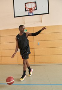 Basketballspiller i en sort ærmeløs trøje og shorts, der udfører en øvelse. Lyse, flerfarvede sneakers og en basketball på banen.