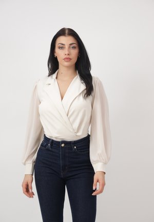Mujer con cabello largo y oscuro, vistiendo una blusa blanca cruzada con mangas largas abullonadas y jeans azul oscuro de cintura alta, sobre un fondo liso.