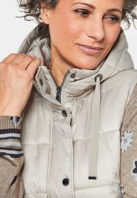 Helle beigen, gesteppte Weste mit hohem Kragen, Reißverschluss und Druckknopfverschlüssen, akzentuiert durch einen Kordelzug, sowie einem strukturierten braunen Pullover mit floralen Mustern.