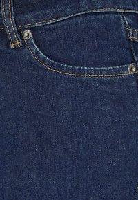 Donkerblauwe denim jeans met een voorzak, koperen stiksels en een metalen knoopdetail. De stof heeft een zachte, lichtjes gestructureerde oppervlakte.