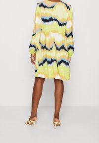 Vestido acampanado de mangas largas con un patrón en zigzag, que presenta colores amarillo, verde, negro y azul, hecho de una tela ligera.