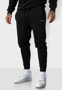 Schwarze Sweatpants aus einem weichen Baumwoll-Mix, mit Seitentaschen, elastischen Bündchen und einem kleinen Logo-Akzent auf der Vorderseite. Kombiniert mit weißen Sneakers.