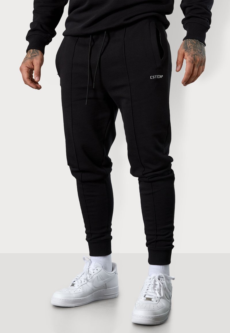 Schwarze Sweatpants aus einem weichen Baumwoll-Mix, mit Seitentaschen, elastischen Bündchen und einem kleinen Logo-Akzent auf der Vorderseite. Kombiniert mit weißen Sneakers.