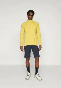 Κίτρινο μακρυμάνικο φανελάκι με φερμουάρ σε συνδυασμό με σκούρες βερμούδες. Διαθέτει ψηλό γιακά και μία τσέπη στην προτομή με φερμουάρ. Τα παπούτσια είναι γκρι με μαύρες λεπτομέρειες.