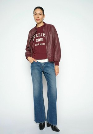 Woman standing wearing maroon leather jacket, maroon sweatshirt with text, blue flared jeans, and black heeled boots against white background.
