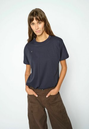 Vrouw met lang bruin haar, draagt een donkerblauw t-shirt met korte mouwen en een bruine broek, met handen in de zakken, staand tegen een effen lichte achtergrond.