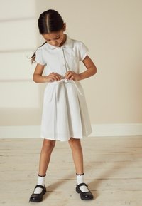 Robe en coton blanche à manches courtes, col et poches. Présente une ceinture nouée et une jupe plissée. Chaussures noires et chaussettes blanches à bords festonnés.