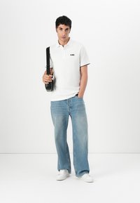 Witte polo met een zwart logo op de borst, gecombineerd met lichtblauwe, wijde jeans en witte sneakers. Een zwarte schoudertas wordt vastgehouden.