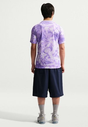 Άνδρας με μωβ κοντομάνικο πουκάμισο με λουλούδια, ναυτικό σκούρο μπλε σορτς με φαρδύ πόδι, γκρι κάλτσες με σχέδια και γκρι αθλητικά παπούτσια, στέκεται με την πλάτη γυρισμένη.