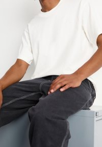 White cotton t-shirt with short sleeves, paired with dark gray jeans featuring contrasting stitching. Model seated on a gray surface.