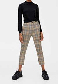 Czarna bluzka z długim rękawem w zestawie z materiałowymi spodniami w kratę w beżowym, czarnym i pomarańczowym wzorze. Modelka ma na sobie masywne czarne sneakersy.