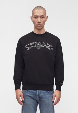 Mand iført sort sweatshirt med teksten "ICEBERG" og en glitrende kaninlogo, kombineret med blå jeans, stående mod en ensfarvet baggrund.