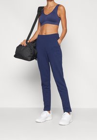 Nainen, joka on pukeutunut tummansiniseen urheilurintaan ja verkkareihin, kantaa mustaa kuntosalilaukkua olkapäällä, jalassaan valkoiset lenkkarit yhtenäisellä taustalla.