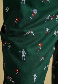 Zaļš audums ar rakstām, kurā redzami mazi, daudzkrāsaini attēli ar regbija spēlētājiem darbībā. Materiāls izceļas ar zigzagveida tekstūru.