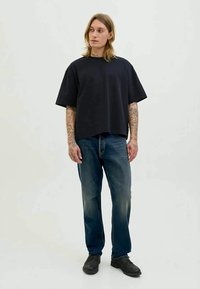 Pessoa com cabelo até aos ombros e tatuagens nos braços, vestindo uma t-shirt preta larga, jeans azuis e sapatos pretos, de pé contra um fundo simples.