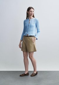 Naine seisab käed taskutes, seljas hele sinine nööpidega kardigan, beežid lühikesed püksid ja leopardimustrilised madalad jalatsid, taust on ühtlane.