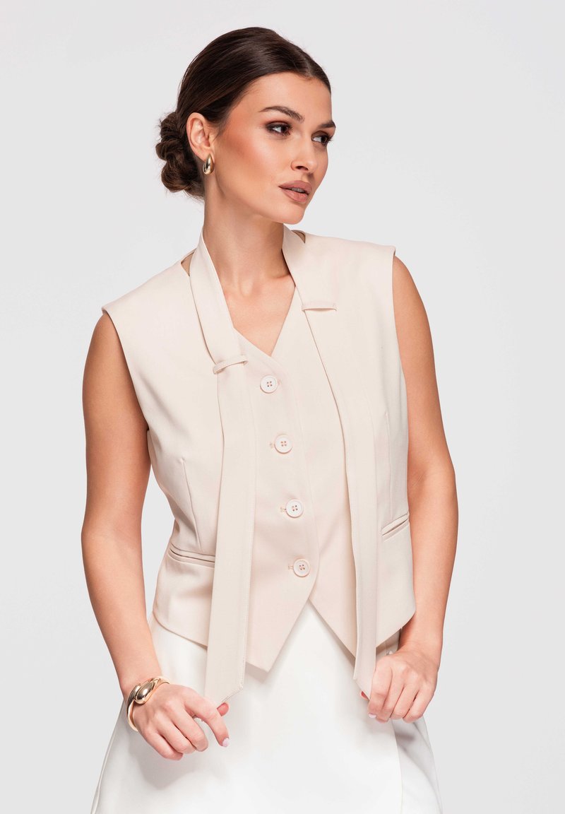 Femme aux cheveux foncés en chignon bas porte un gilet beige sans manches avec des boutons et des boucles d'oreilles créoles dorées, tenant les liens du gilet devant un fond uni.